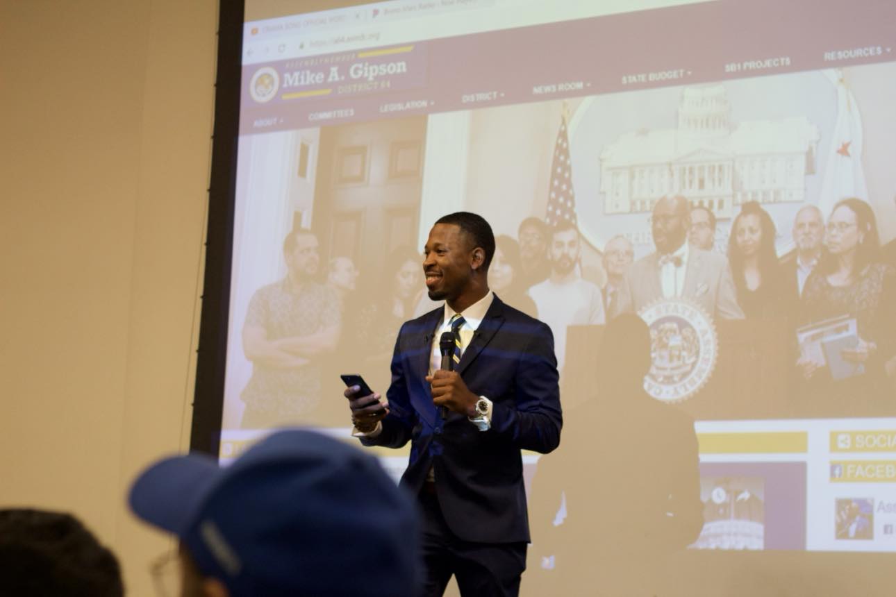 Brian E. Sibley on stage with a microphone introducing Assemblyman Mike A. Gipson at Cal State Dominguez Hills